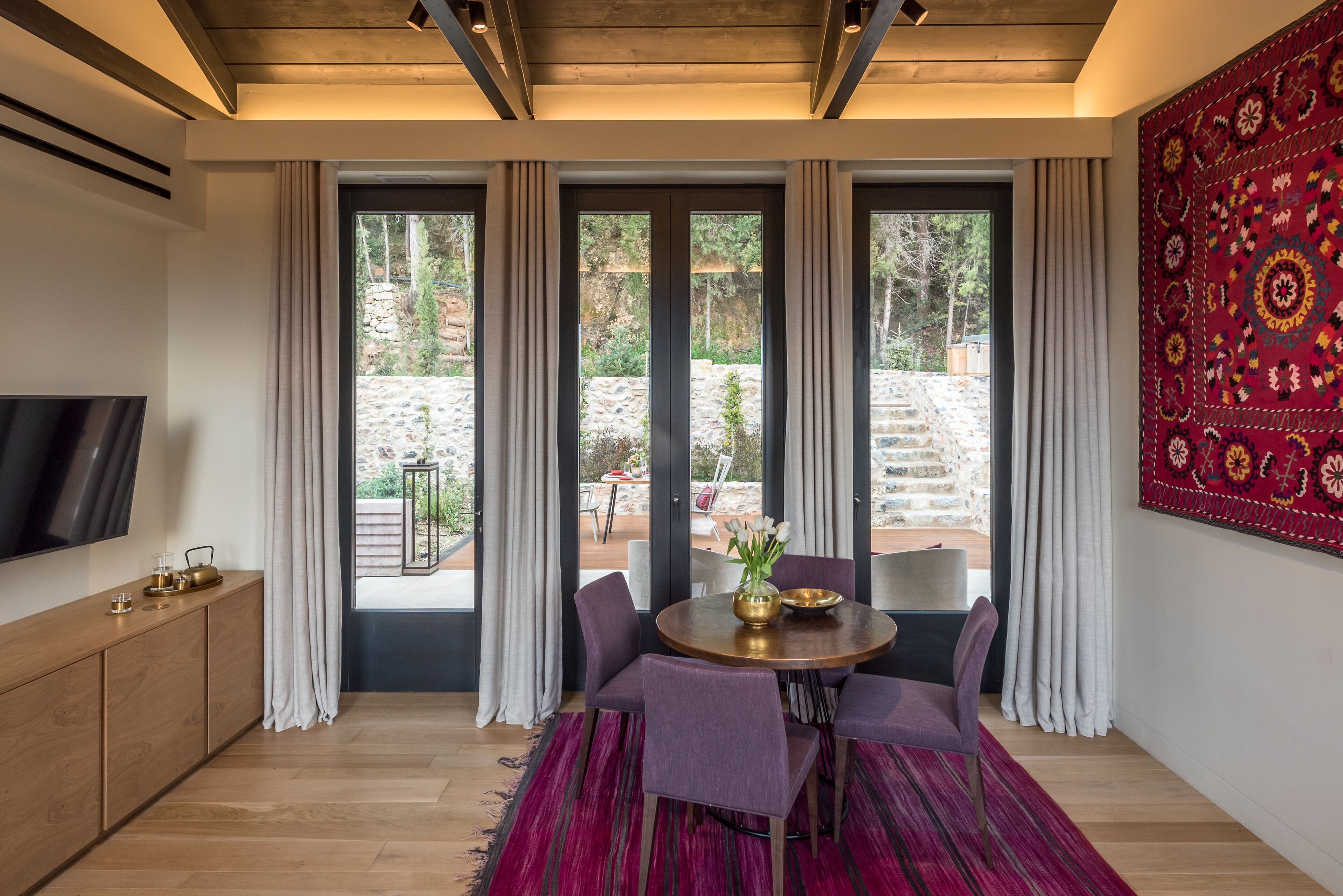 Una habitación moderna con techo de madera, televisor a la izquierda, una mesa de comedor con sillas y vistas a un patio de piedra.
