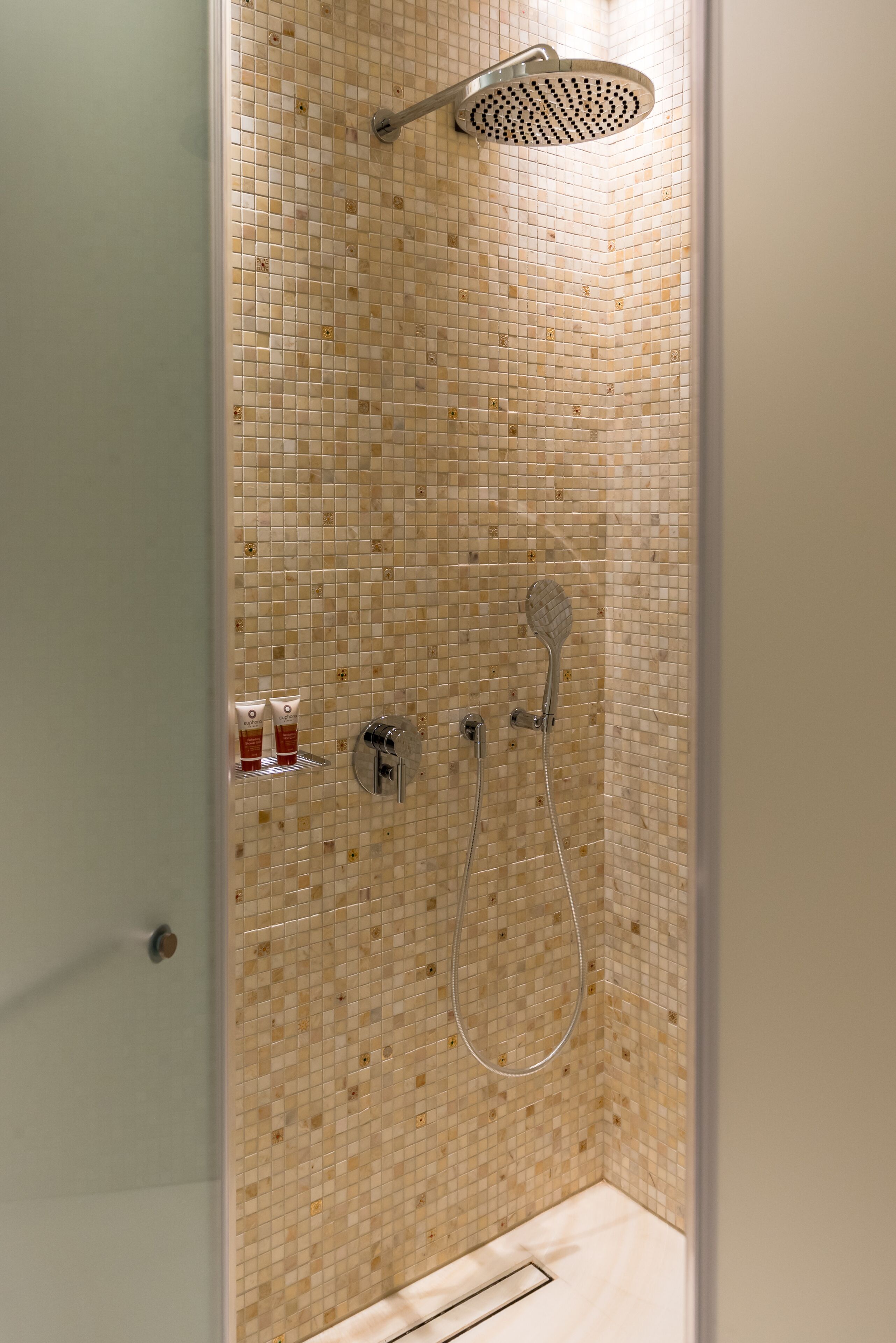 A shower with a mosaic tile wall, a handheld showerhead, and a wall-mounted showerhead.