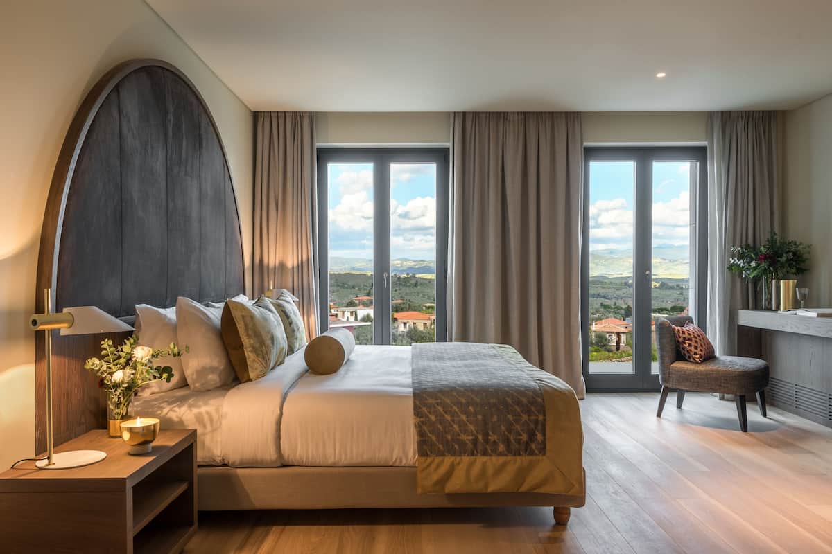 Une chambre à coucher avec un grand lit, une table de chevet, une chaise et une vue sur la campagne, grâce à de grandes fenêtres.