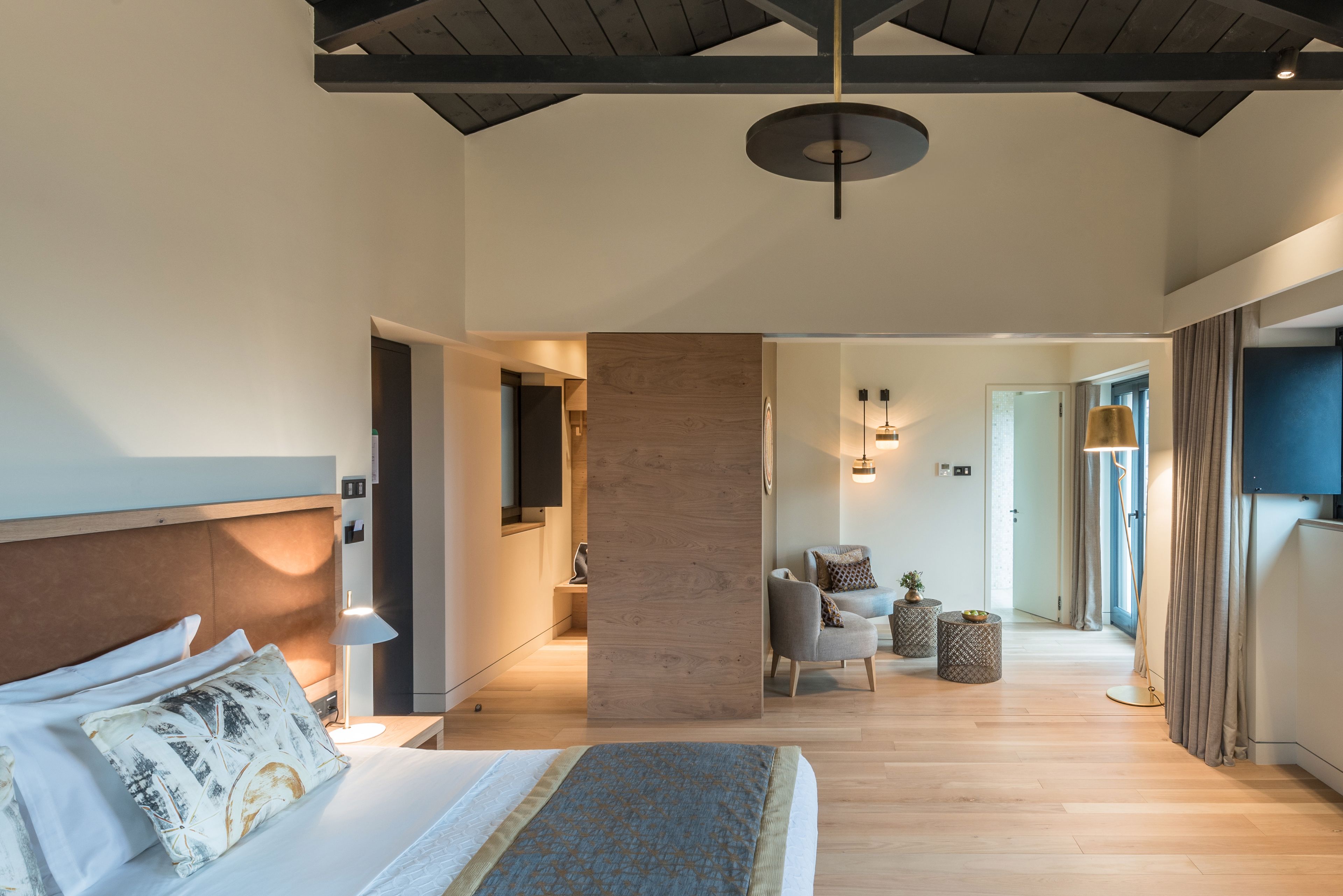 A modern hotel room with a large bed, a wooden accent wall, a seating area with a chair and side table, and a view of the hallway.