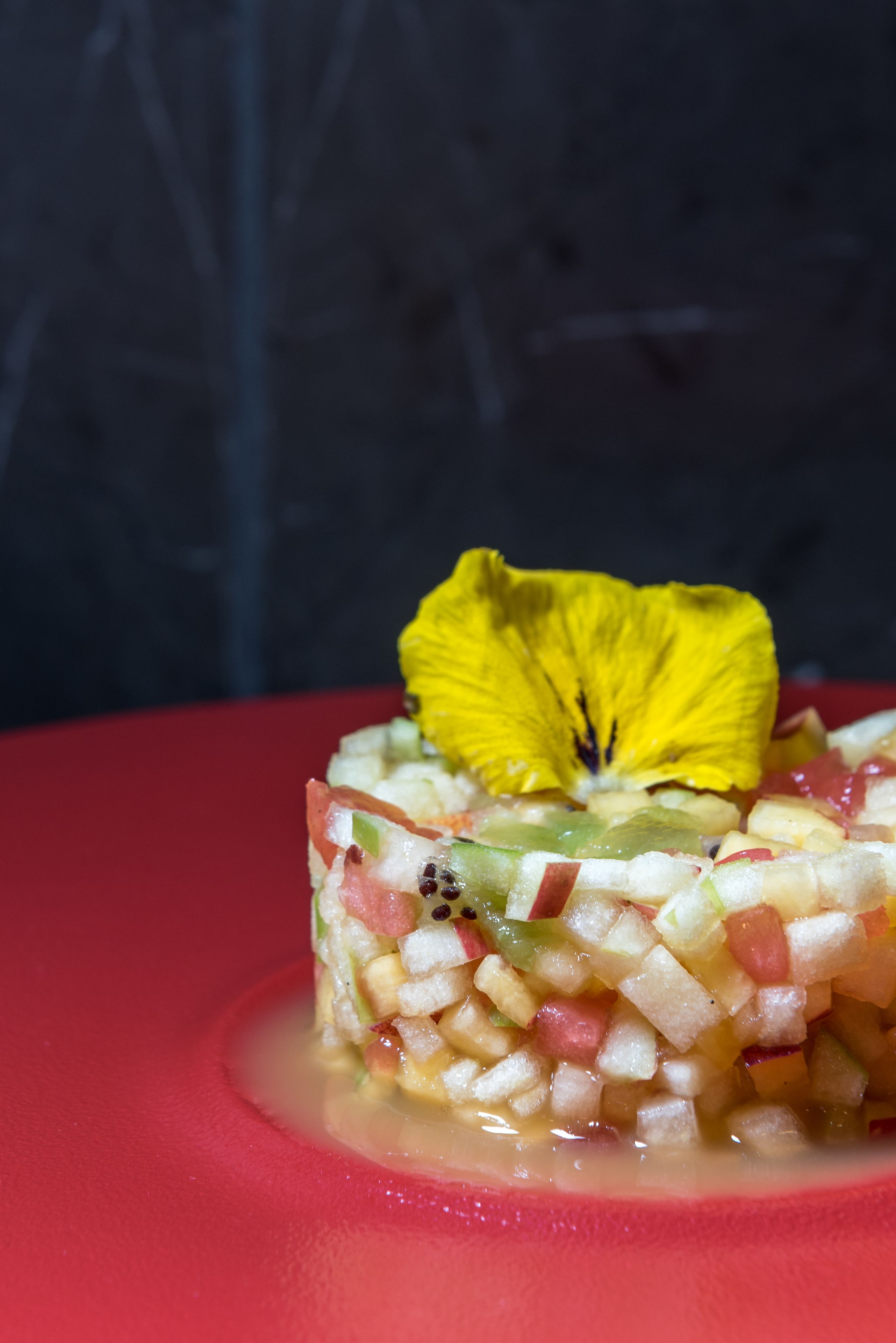 A dessert with diced fruits and a yellow flower on a red plate.