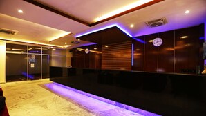 A modern hotel lobby with a marble reception desk, a clock on the wall, and a glass door leading to the rooms.