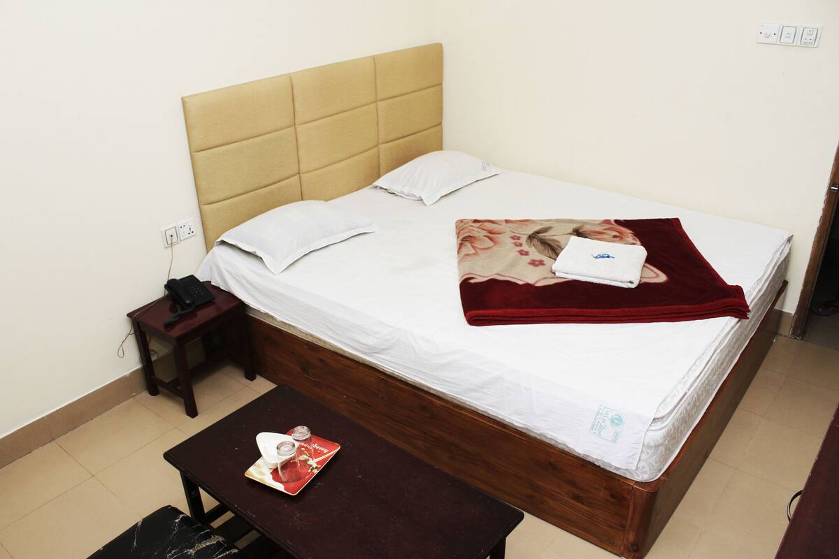 Ein Einzelbett mit einem Holz-Kopfstück, weißer Bettwäsche und einer roten Decke. Ein kleiner Nachttisch mit Telefon und Zeitschrift steht daneben. Vor dem Bett steht ein Holztisch mit einer Tablett voller Gegenstände.