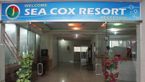 L'entrée de l'aire d'accueil du Sea Cox Resort se distingue par une porte en verre, un comptoir d'accueil et un panneau d'affichage.