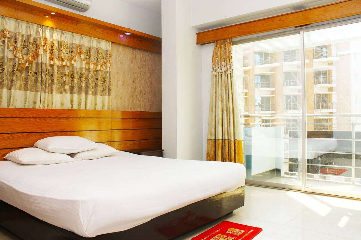 Une chambre d’hôtel avec un lit, une tête de lit en bois et une vue sur un immeuble, visible à travers une porte vitrée.
