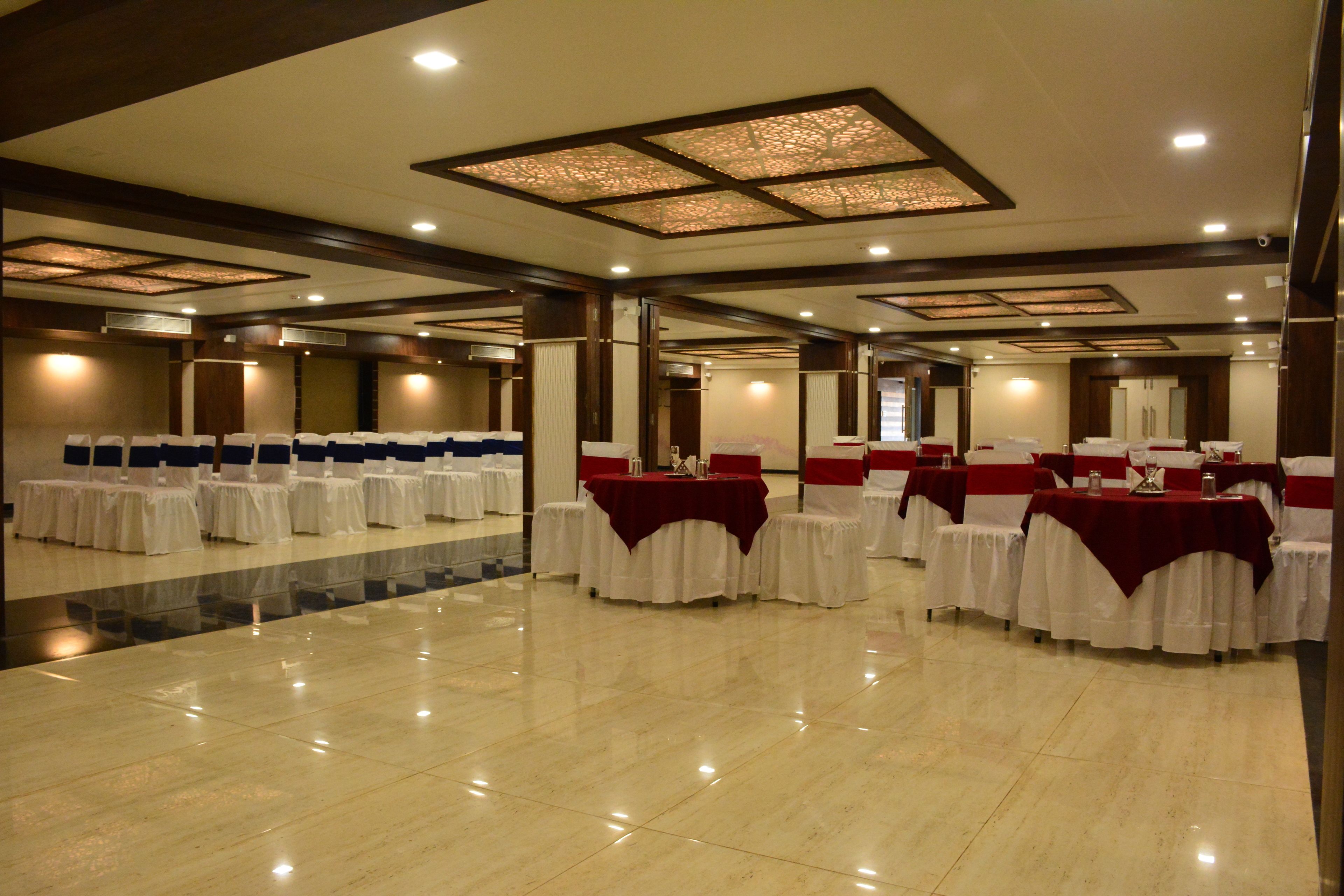 Una espaciosa sala de banquetes con múltiples mesas redondas, cada una cubierta con manteles blancos y fajas rojas. La sala cuenta con un suelo de mármol pulido y un techo alto con iluminación empotrada.