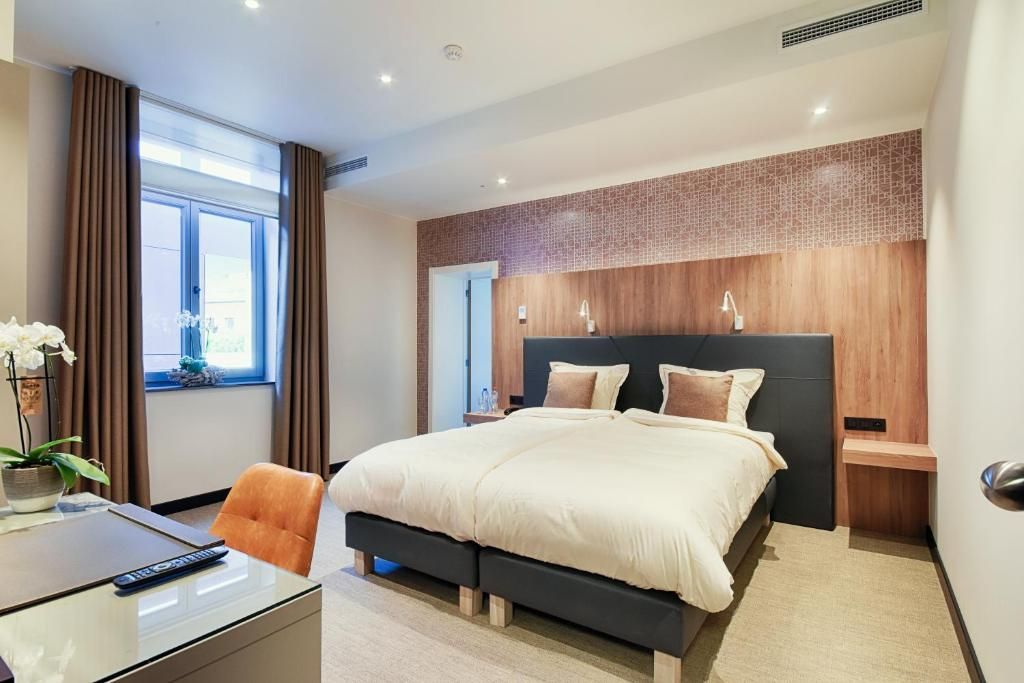 Una habitación de hotel moderna con una cama grande, un escritorio con silla, una ventana con cortinas y una planta decorativa.