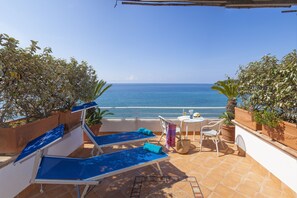 Terraço com duas espreguiçadeiras, uma mesa pequena e cadeiras, com vista para o mar.