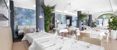 Un restaurante moderno con mesas blancas, sillas color beige y un mural azul en la pared.