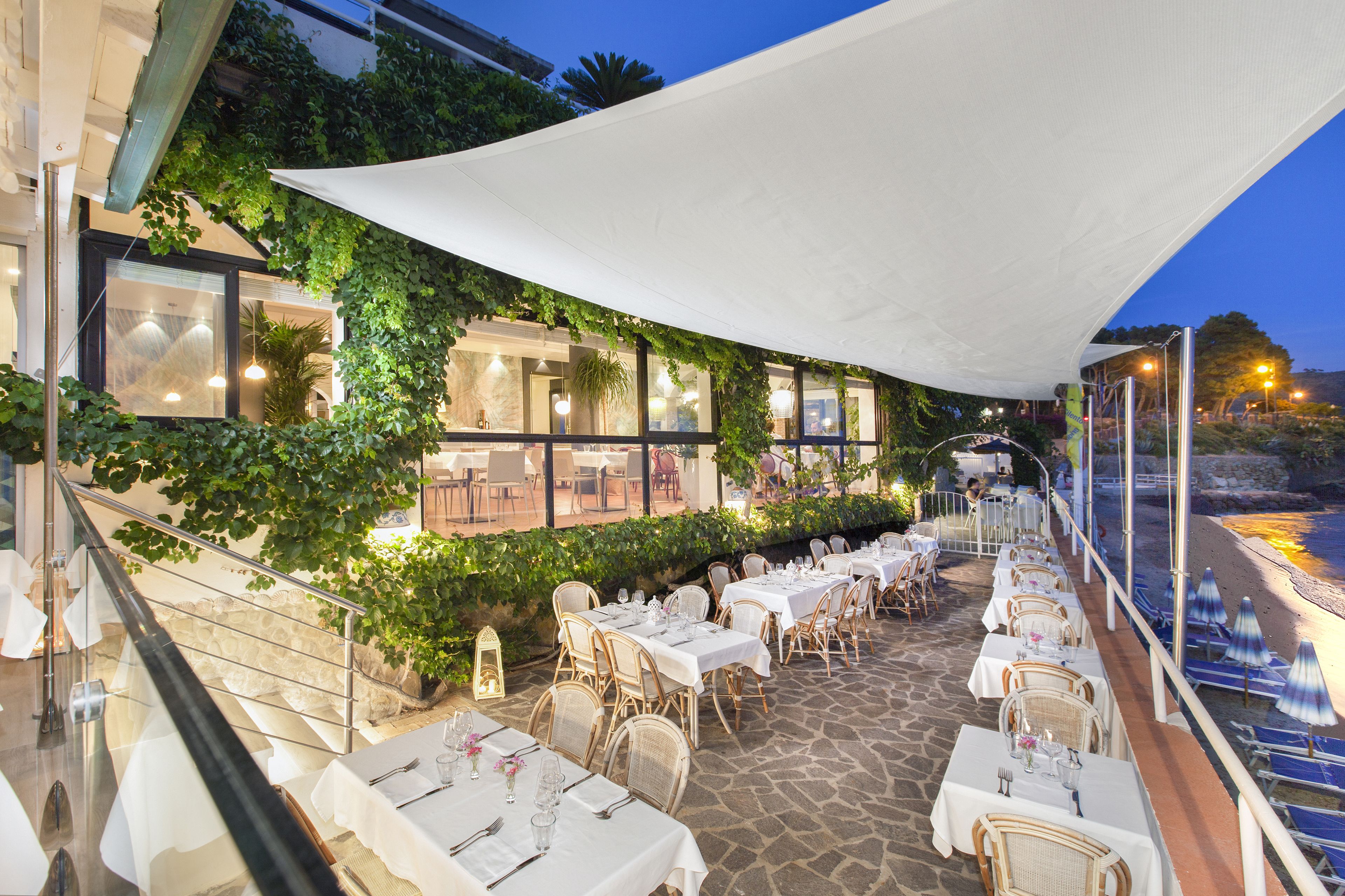 Un restaurante al aire libre con mesas blancas y sillas de mimbre, frente a un entorno verde y vistas al mar.