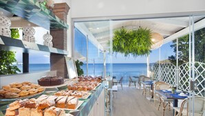 A buffet with various pastries and cakes, a seating area with tables and chairs, and a view of the ocean through large windows. - Hotel Il Cefalo (Castellabate)