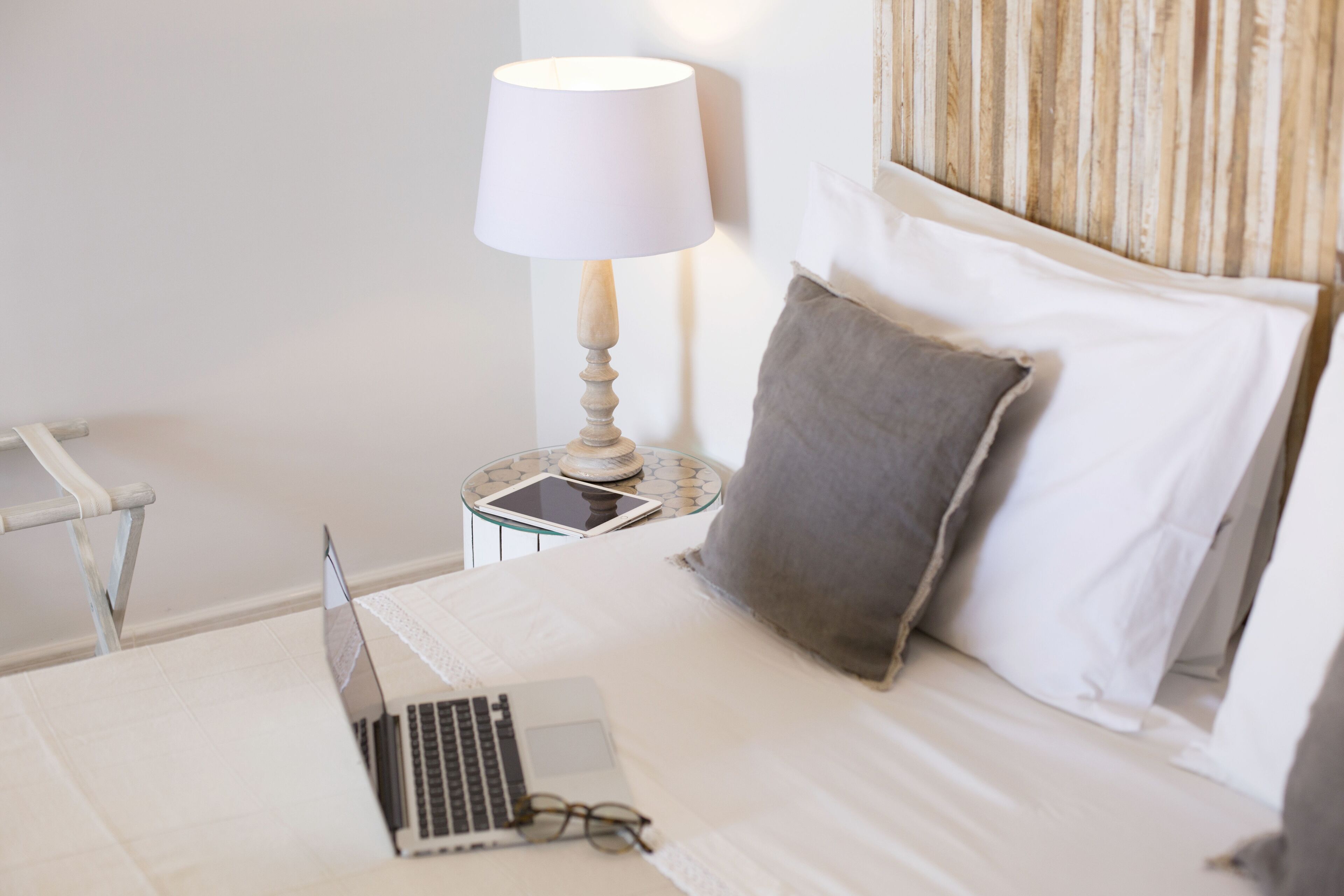 A neatly made bed with a laptop, a lamp, and a tablet on a bedside table.