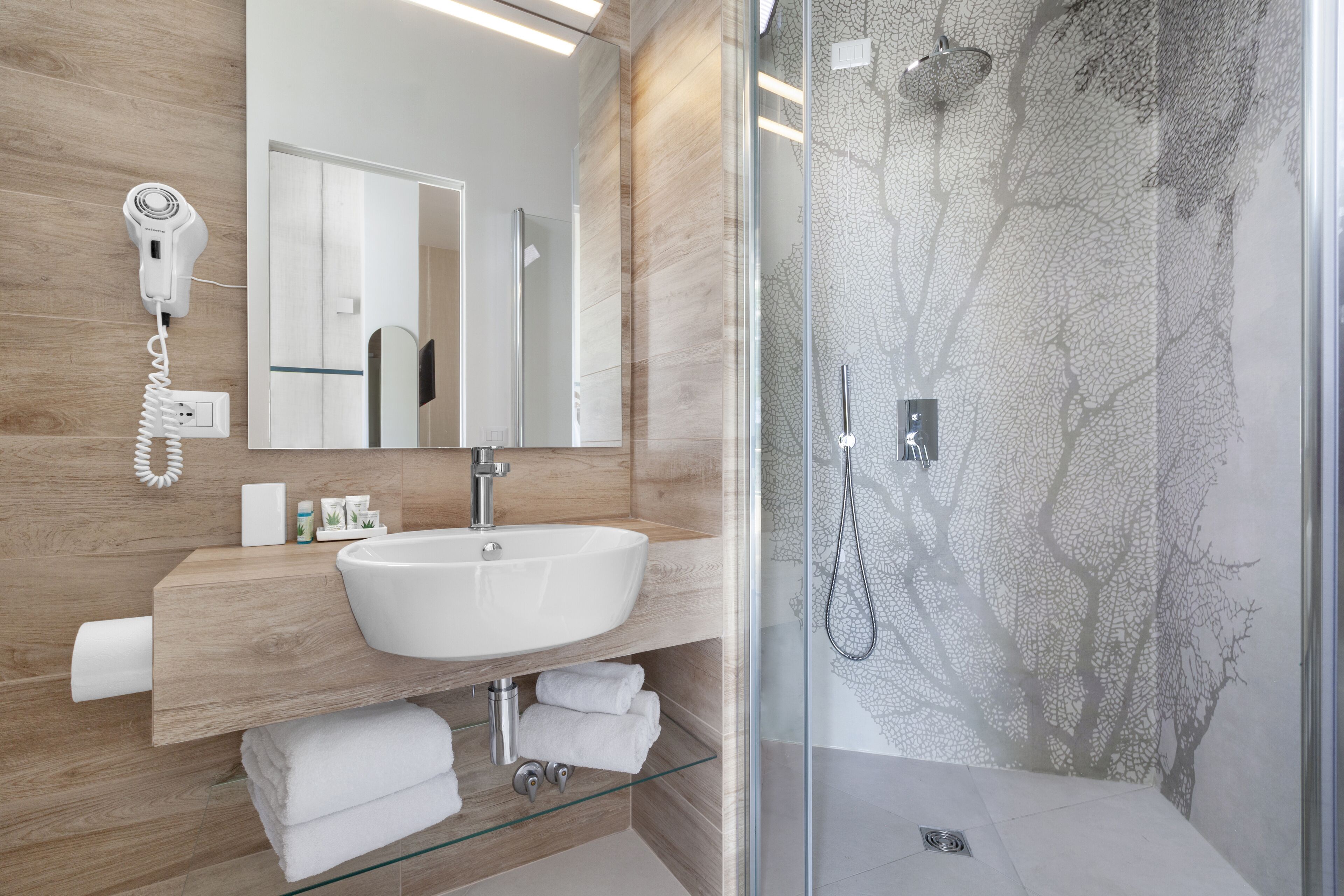 A modern bathroom with a wooden vanity, a white sink, a glass shower enclosure, and a hairdryer on the wall.
