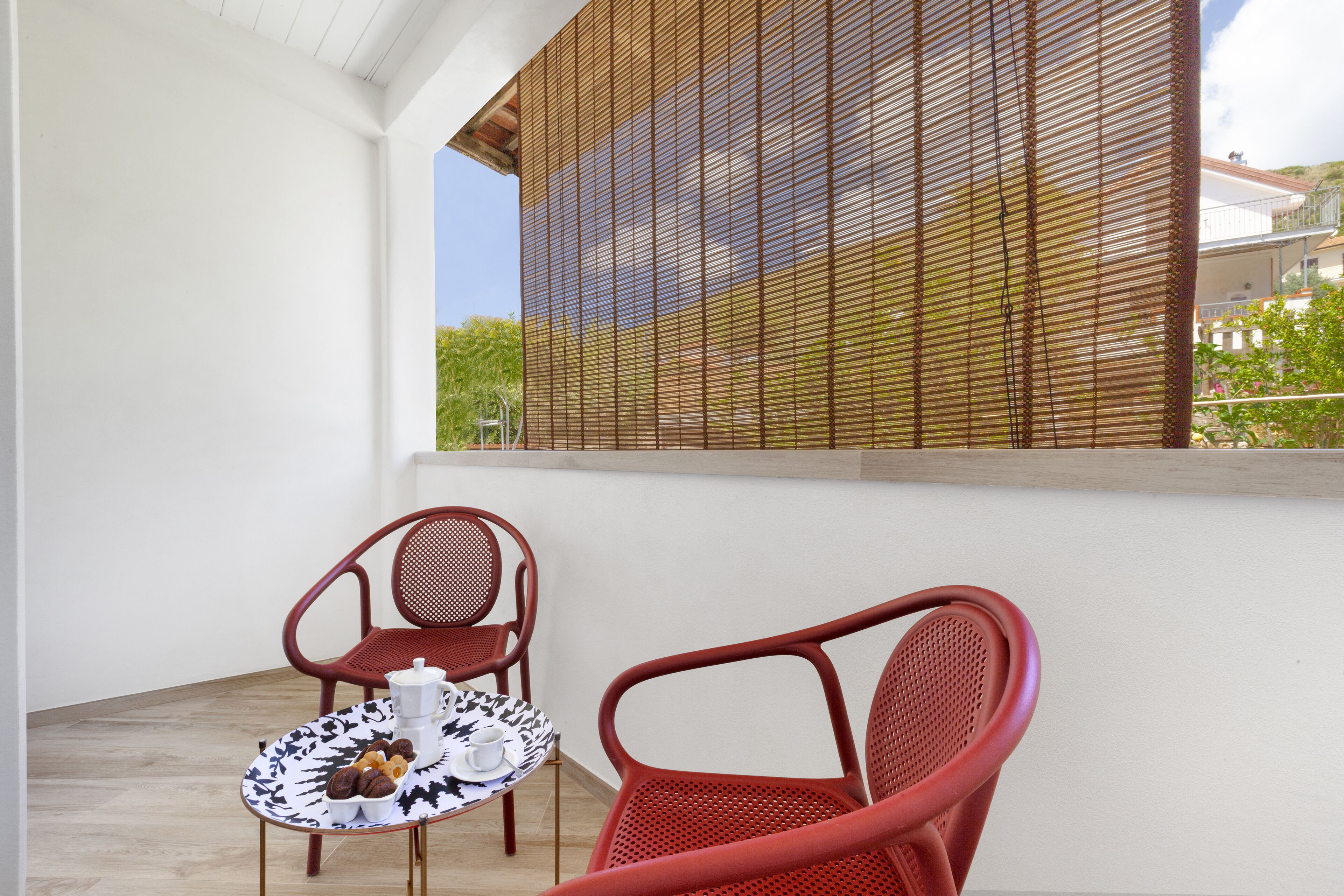 Un balcon avec deux chaises rouges et une petite table recouverte d’une nappe à motifs noirs et blancs, un pot blanc et une tasse.