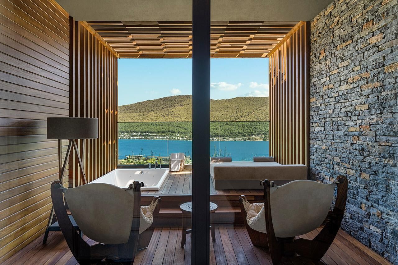 Ein Balkon mit Blick auf einen See und Hügel, Holzboden, zwei gepolsterte Stühle, ein kleiner Tisch und eine Stehlampe.