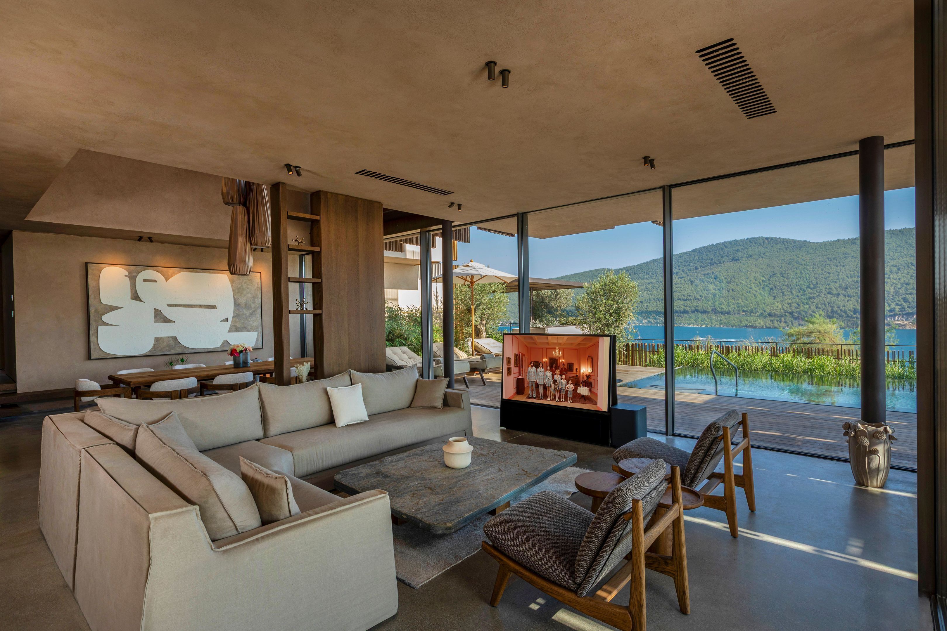 A modern living room with a large sofa, a coffee table, and a television. The room has large windows with a view of a lake and mountains.