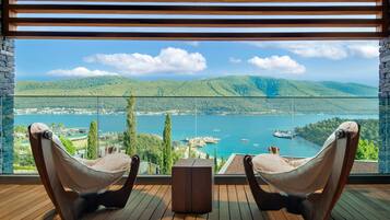A balcony with two cushioned chairs and a small table, overlooking a lake with boats and a dock, surrounded by greenery and hills.
