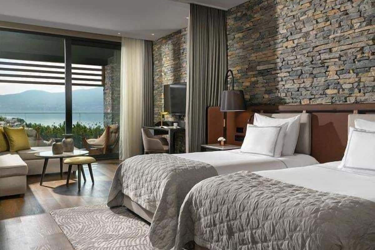 Una habitación de hotel moderna con dos camas, un escritorio y una silla. La habitación cuenta con una pared de piedra y un ventanal con vistas a las montañas.