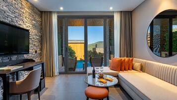 A modern living room with a stone wall, a flat-screen TV, a sofa with orange cushions, a dining table with a wine glass, and a view of a pool and hills.