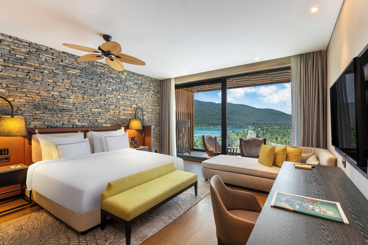 Una habitación de hotel moderna con una cama grande, un sofá, una mesa de comedor y un balcón con vistas a las montañas.