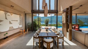 A modern dining area with a long table and chairs, large windows with a view of a lake and mountains, and a large abstract art piece on the wall.