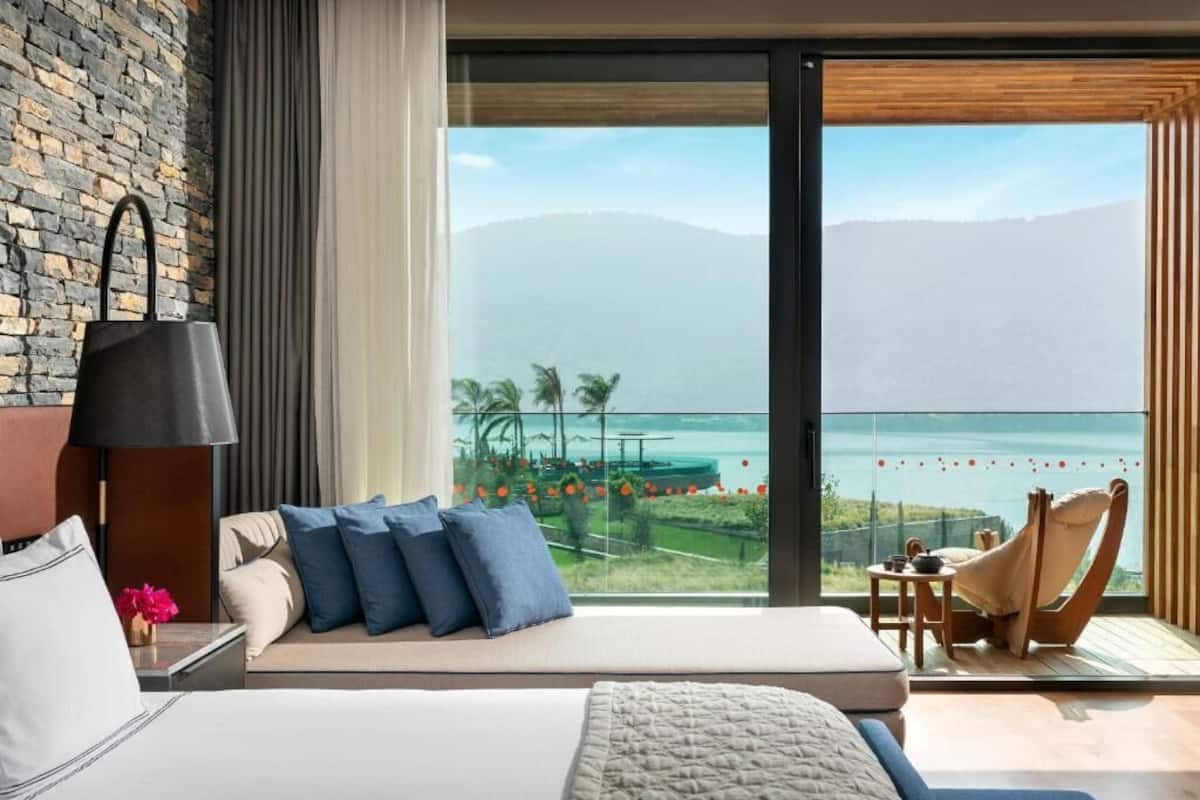 Habitación de hotel moderna con una cama grande, vista al mar, un área de descanso con una silla y una pared de ladrillo.