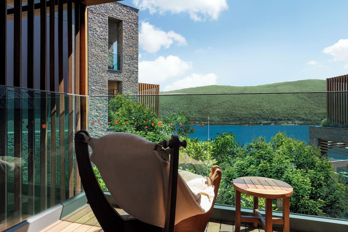 Un balcón con una silla y una mesita, con vista a un paisaje pintoresco de un lago y colinas.