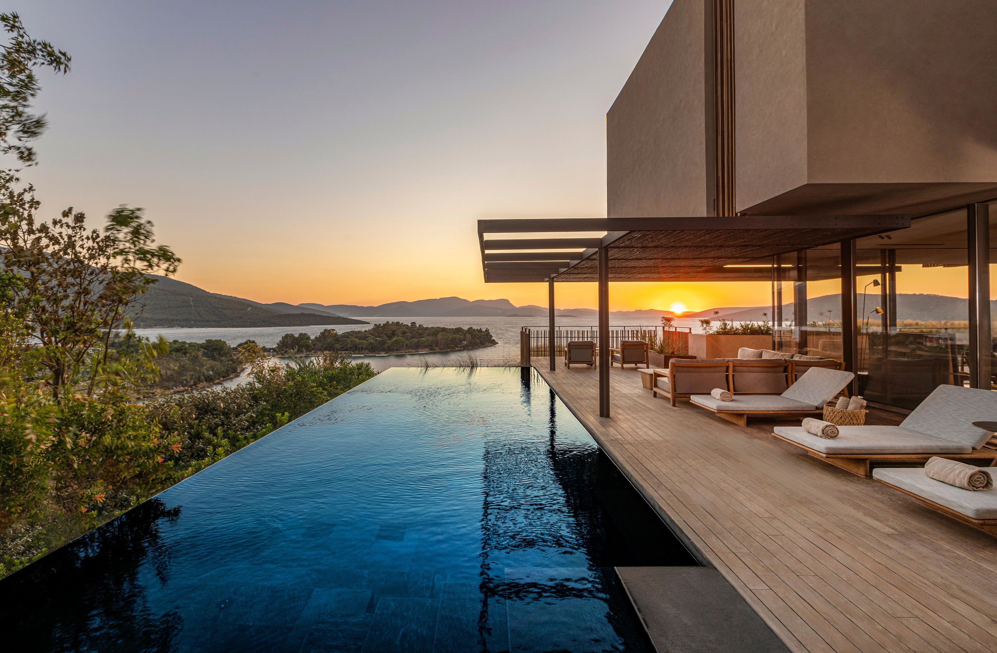 Une piscine à débordement moderne, dotée d’une terrasse en bois et de chaises longues, offrant une vue imprenable sur un magnifique coucher de soleil.