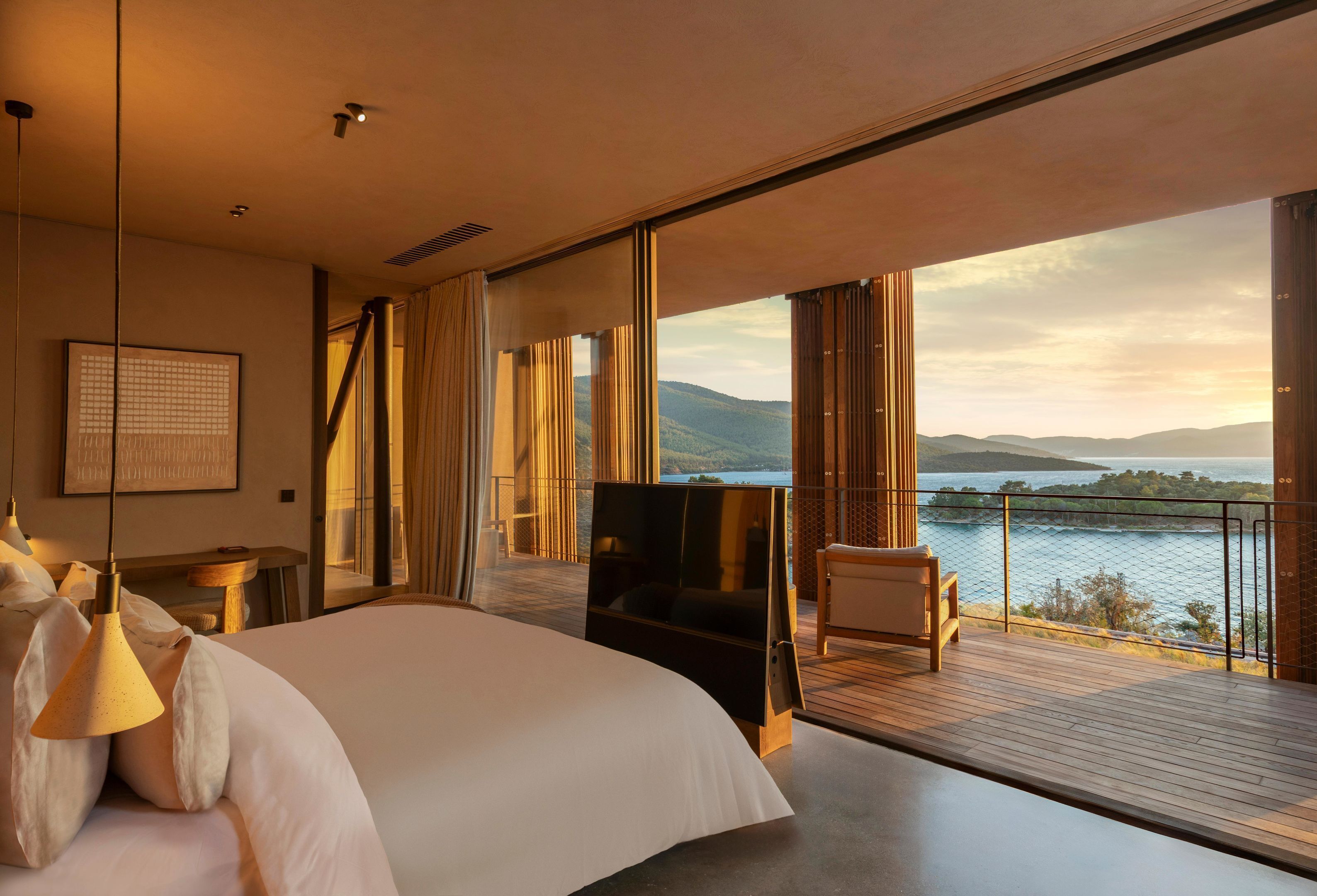 Habitación de hotel moderna con una cama grande, televisión y balcón con vistas al agua y a las montañas.