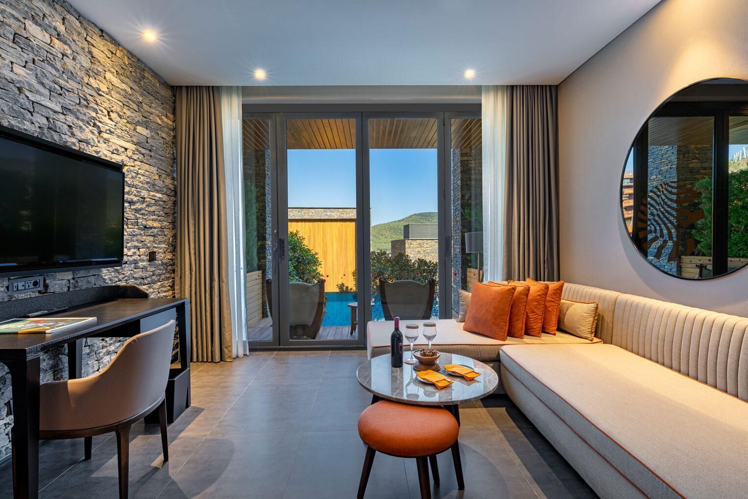 A modern living room with a stone wall, a flat-screen TV, a sofa with orange cushions, a dining table with a bottle and glasses, and a view of a pool and mountains.