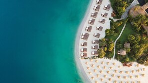 Vue aérienne d’un complexe hôtelier en bord de mer, avec des rangées de transats et de parasols, une plage de sable et une mer d’un bleu limpide.
