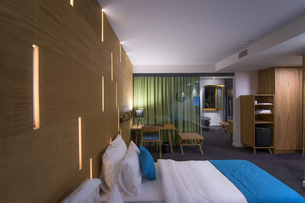 A hotel room with a wooden headboard, a bed with white and blue bedding, a desk with a chair, a small table, and a bookshelf.
