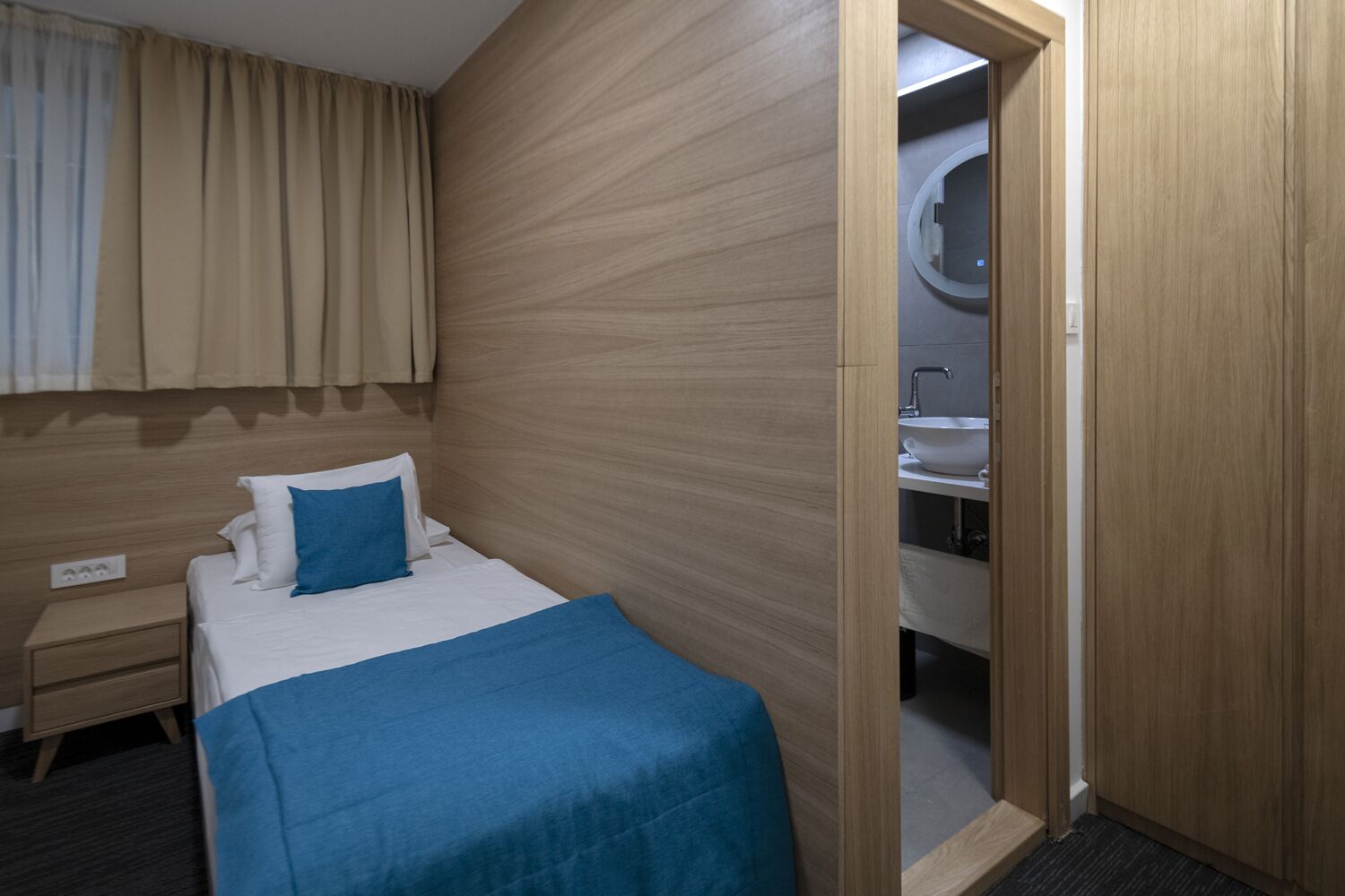 A hotel room with a bed, a nightstand, a wooden headboard, and a bathroom visible through an open door.