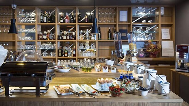 A buffet setup with various food items, a coffee machine, and a wine and glass display in the background. - Hotel Sana (Sarajevo)