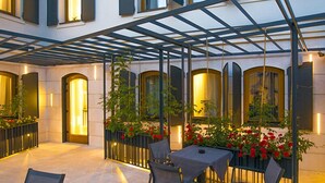 A covered outdoor seating area with tables and chairs, surrounded by windows with curtains, and plants in black metal planters. - Hotel Sana (Sarajevo)