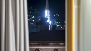 A view through a window with a curtain, featuring a building with a spire and lights, and a detailed architectural facade with arched windows and decorative elements. - Hotel Sana (Sarajevo)