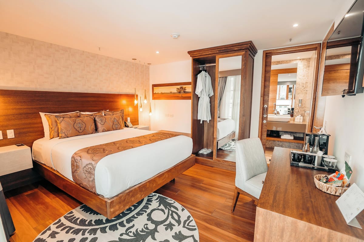 A modern hotel room with a large bed, wooden headboard, a desk with a chair, a mirror, and a small kitchen area.