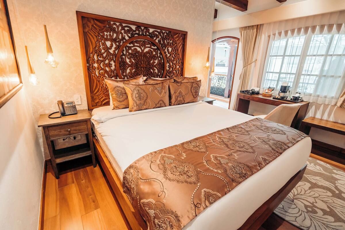 A hotel room with a large bed, a wooden headboard, a desk with a chair, and a view of the outside through a window.