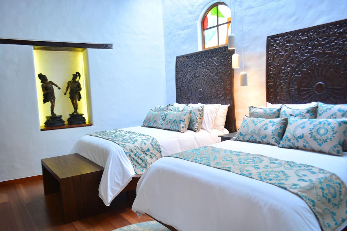 A bedroom with two beds, a wooden bench, a wall-mounted shelf with sculptures, and a stained glass window.