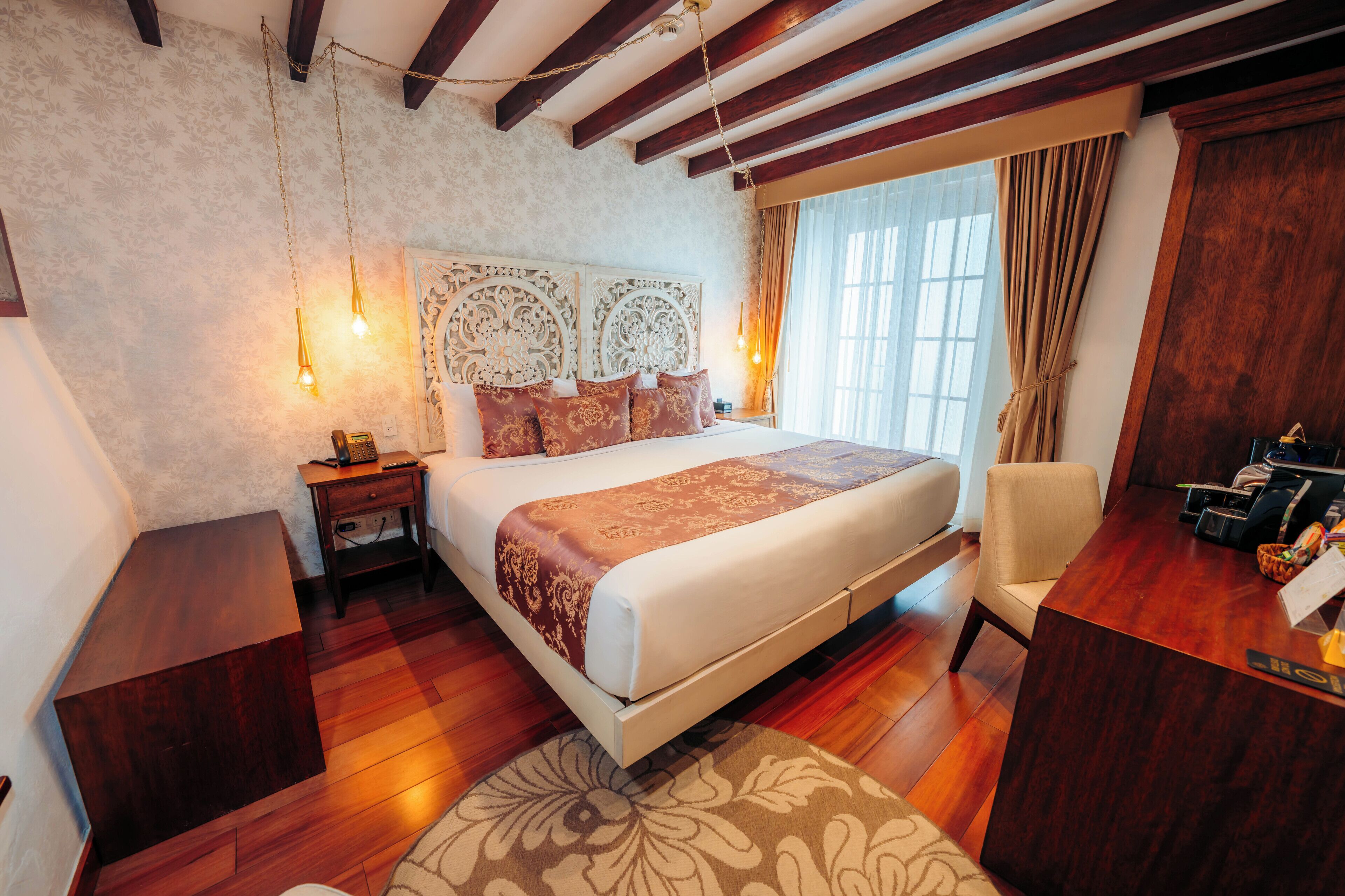 A hotel room with a large bed, wooden flooring, a desk with a chair, and a window with curtains.