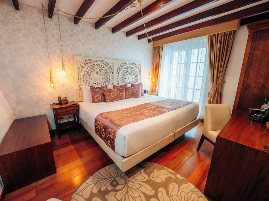 Habitación de hotel con una cama grande, piso de madera, un escritorio con silla y una ventana con cortinas.