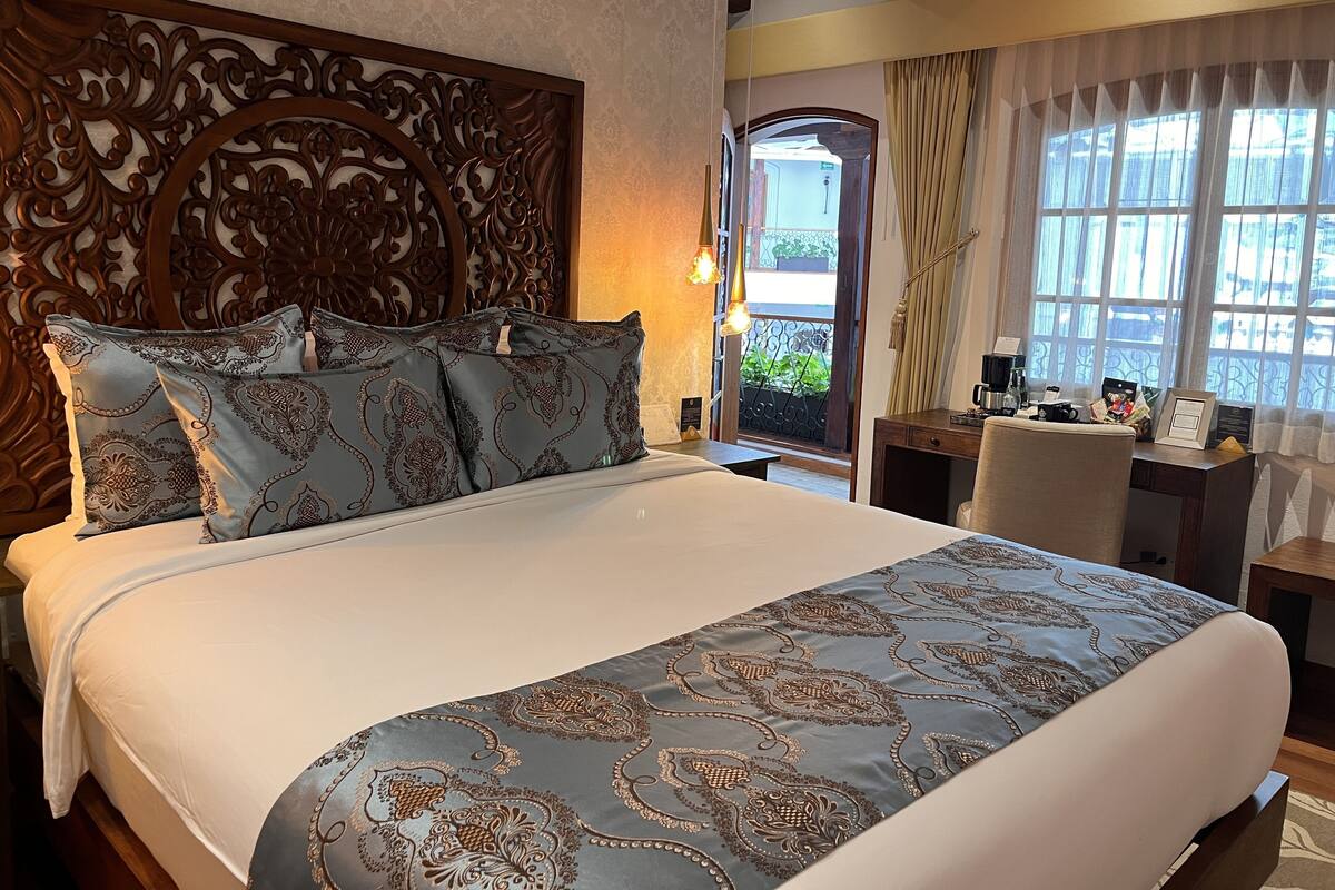 A hotel room with a large bed, a wooden headboard, a desk with a chair, and a view through a window.