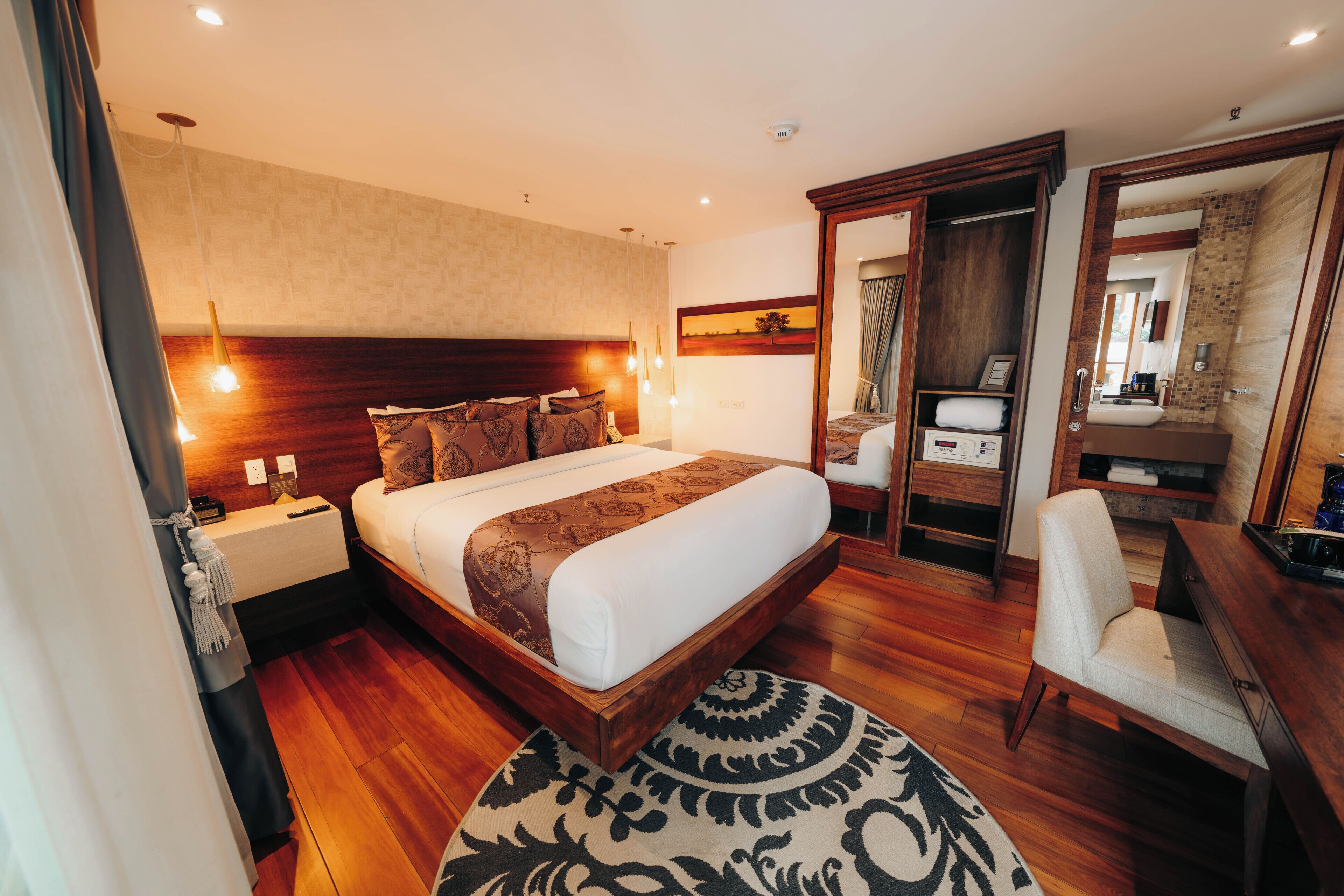 Habitación de hotel con una cama grande, mesitas de noche, un escritorio con silla, un espejo y un baño visible a través de una puerta abierta.