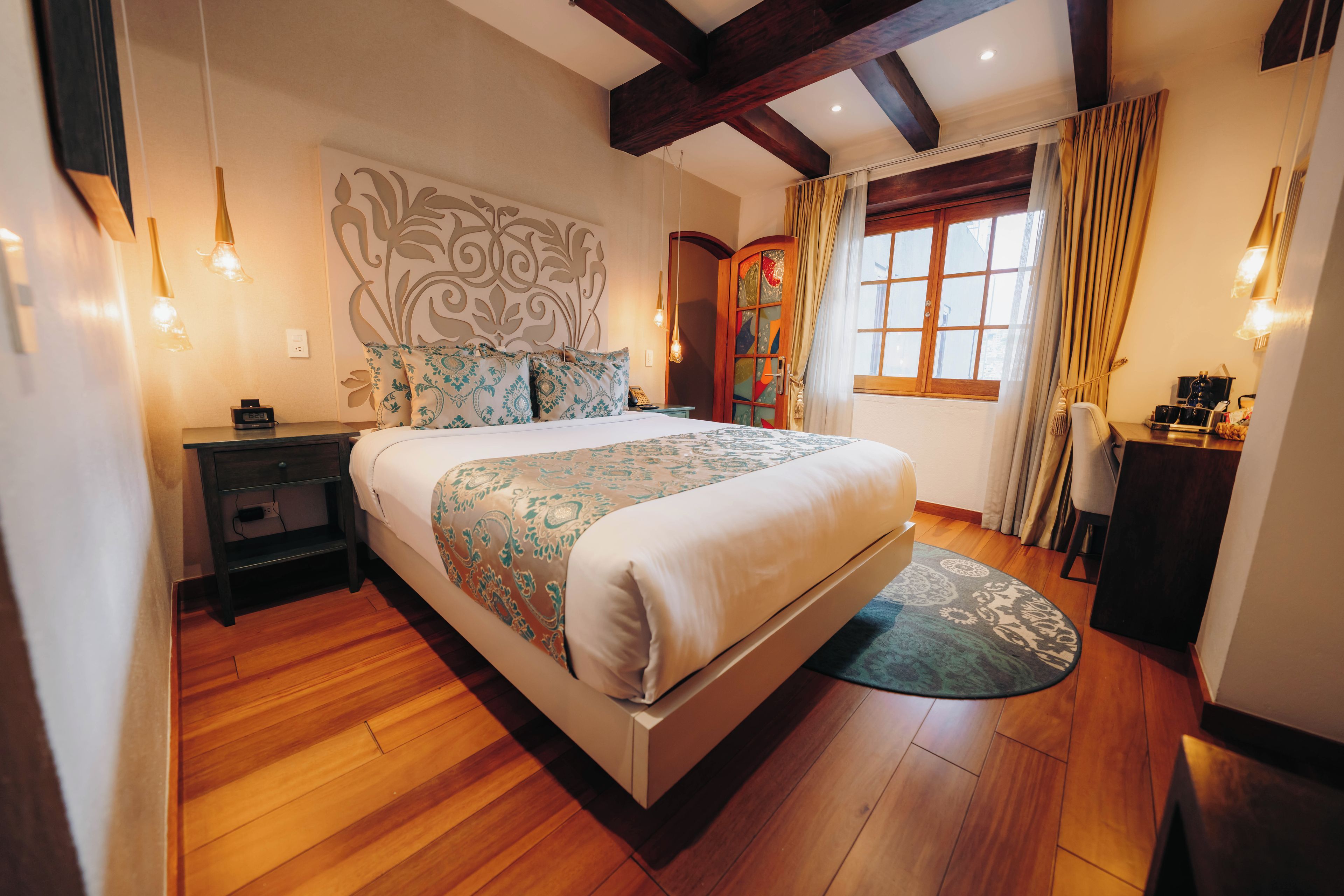 Un dormitorio con una cama grande, piso de madera, un cabecero decorativo, lámparas de noche y una ventana con cortinas.