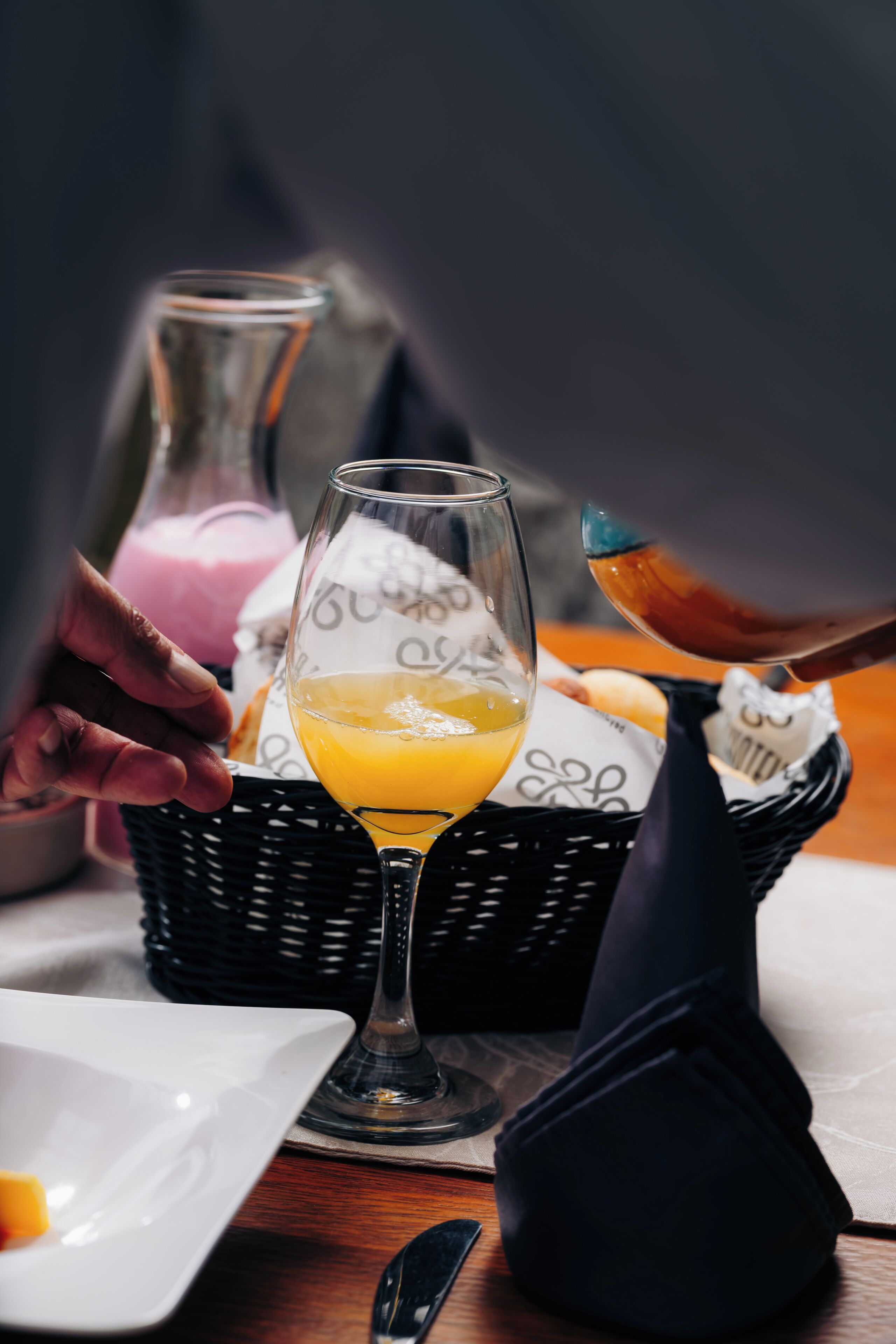 Et spisebord med et glas appelsinjuice, en kurv med brød og en flaske lys rosa drik.