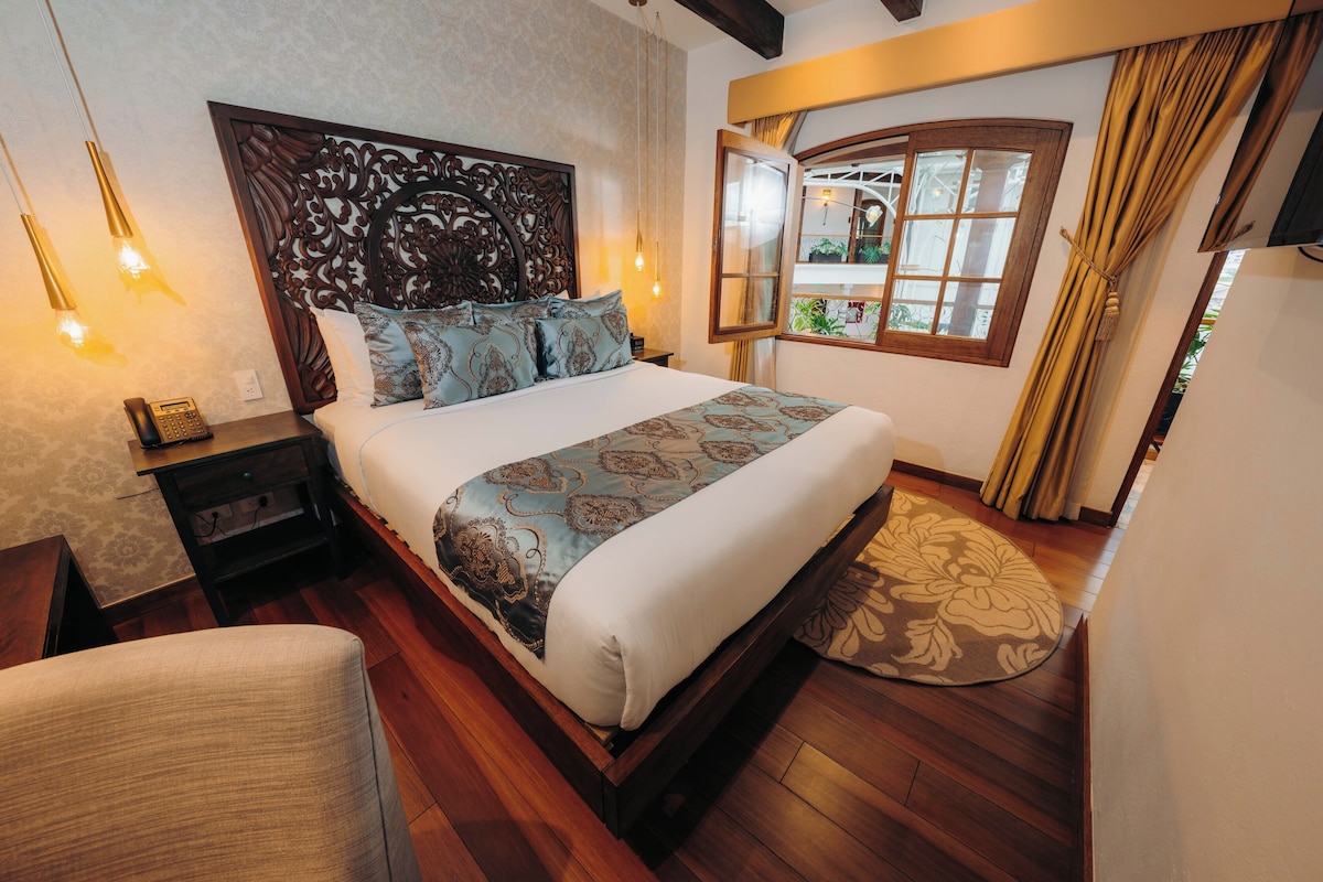 A hotel room with a large bed, a wooden headboard, a bedside table with a telephone, and a window with curtains.