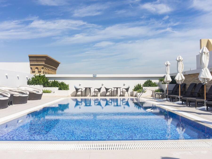 A rooftop pool with lounge chairs and umbrellas, a clear sky, and a building in the background.