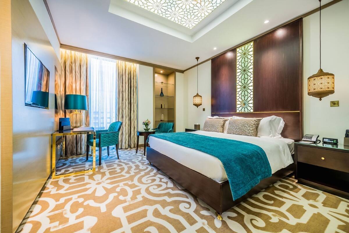 A hotel room with a large bed, a desk, two chairs, a TV, and a decorative ceiling light.