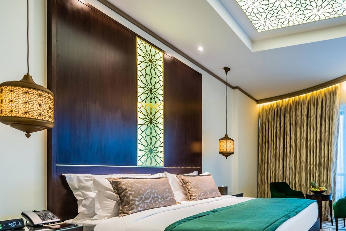 A hotel room with a large bed, a nightstand with a phone, a decorative wall panel, a ceiling light, and a patterned carpet.
