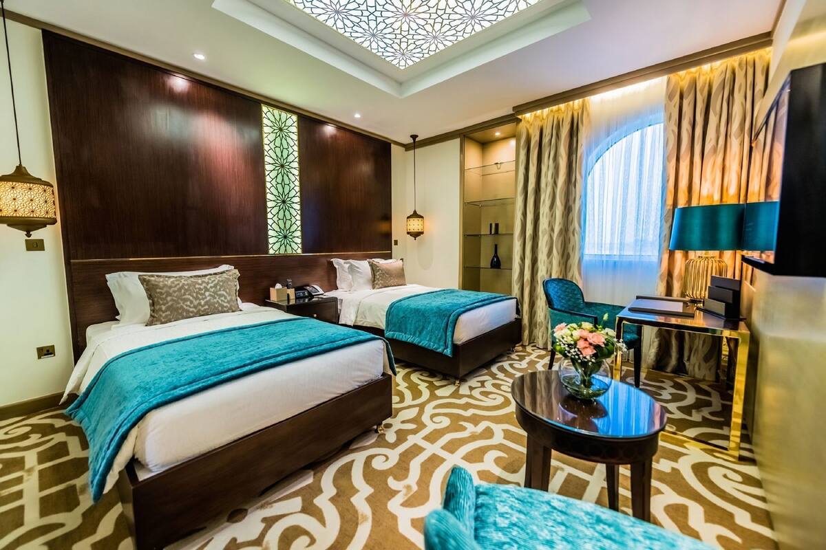 A hotel room with two beds, a desk, a chair, a decorative ceiling, and a patterned carpet.